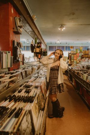 Woman in a cardigan, plaid dress and boots listens to headphones while browsing crates of vinyl records in a record shop.