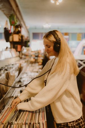Blonde woman in a cream knit sweater and headphones flips through vinyl records in a bin at a vintage record shop.
