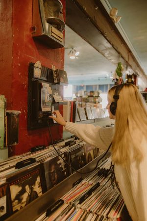 Woman with long blonde hair and headphones listens to a record at a listening station in a cozy vinyl record store.