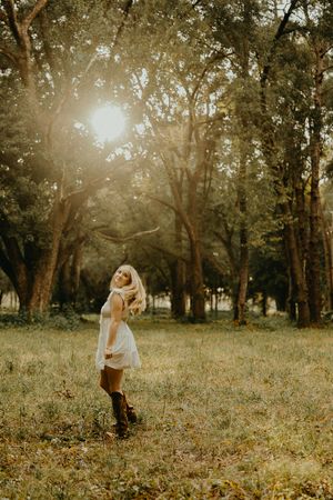 Blonde woman in a white dress and brown boots twirls in a grassy field beneath tall trees with golden sunlight filtering through.