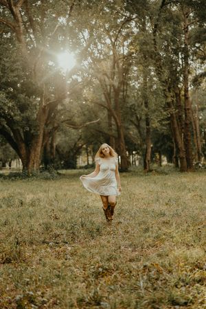 Woman in a white dress and cowboy boots walks through a grassy field as golden sunlight filters through oak trees.