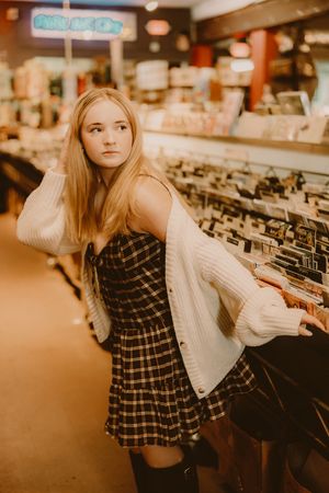 Young woman in a plaid dress and white cardigan looks over her shoulder while browsing vinyl records in a music store.