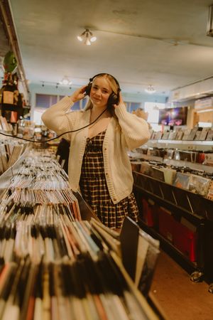 Woman in a cream cardigan and plaid dress wears headphones while browsing vinyl records in a cozy record store.