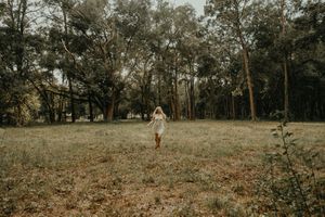 Woman in a white dress walks across a grassy field surrounded by tall trees in soft natural light.