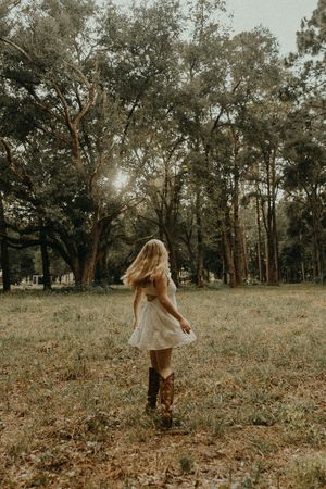 Woman in a white dress and brown cowboy boots twirls in a sunlit grassy field beneath tall oak trees.