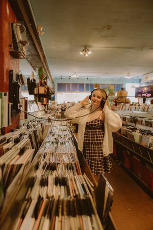 Woman in a plaid dress and white cardigan listens to headphones while browsing rows of vinyl records in a cozy record store.
