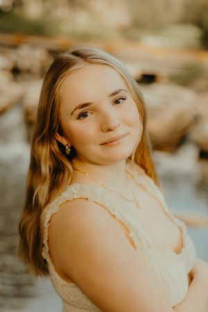 Smiling young woman with long blonde hair in a cream lace top poses beside a sunlit rocky stream.