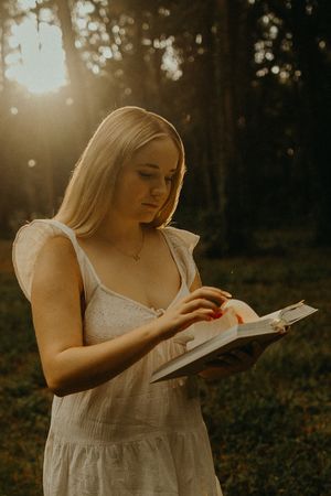 Blonde woman in a white sundress reads an open book outdoors in warm golden-hour light among the trees.