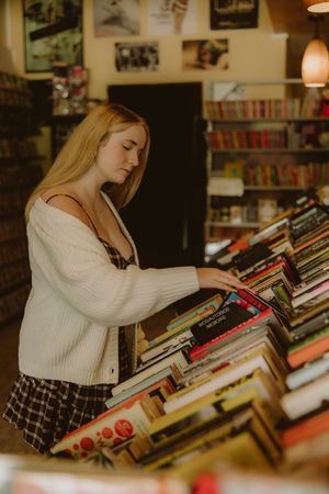 Young woman with blonde hair in a white cardigan and plaid skirt browses stacks of vinyl records in a cozy record store.