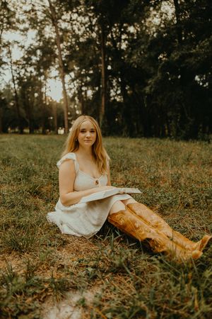 Woman in a white dress and tan cowboy boots sits on green grass holding an open book in a wooded park.