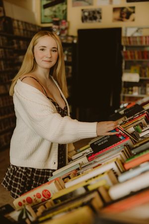 Young woman with long blonde hair in a white cardigan and plaid skirt browses stacks of books in a cozy used bookstore.