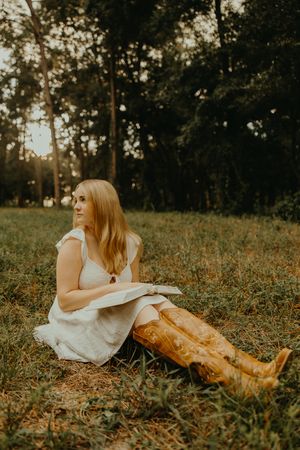 Woman in a white dress and tan cowboy boots sits in a grassy field with an open book, gazing off beneath tall trees.