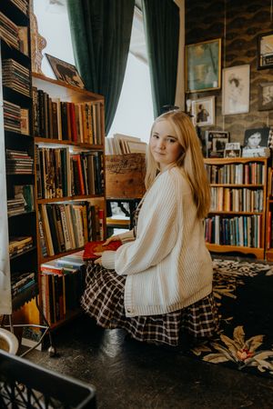 Woman with long blonde hair in a cream cardigan and plaid skirt sits holding a red book in a cozy vintage bookstore.