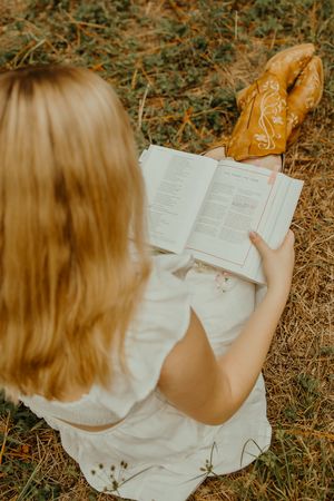 Woman in a white dress sits on dry grass reading an open book, with tan embroidered cowboy boots resting nearby.