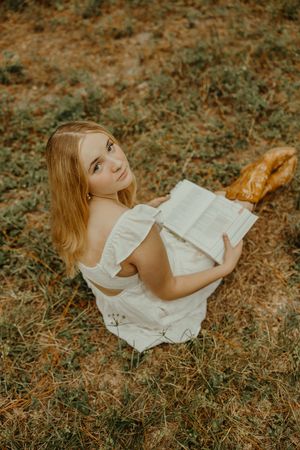 Overhead view of a woman in a white dress sitting on grass, holding an open book and looking up at the camera.