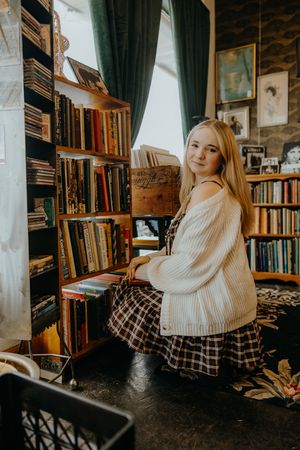 Smiling blonde woman in a cream knit cardigan and plaid skirt sits beside wooden bookshelves in a charming secondhand bookshop.