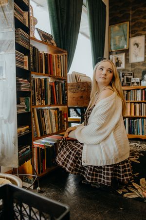 Woman with long blonde hair in a cream cardigan and plaid skirt sits beside crowded wooden bookshelves in a cozy vintage bookstore.
