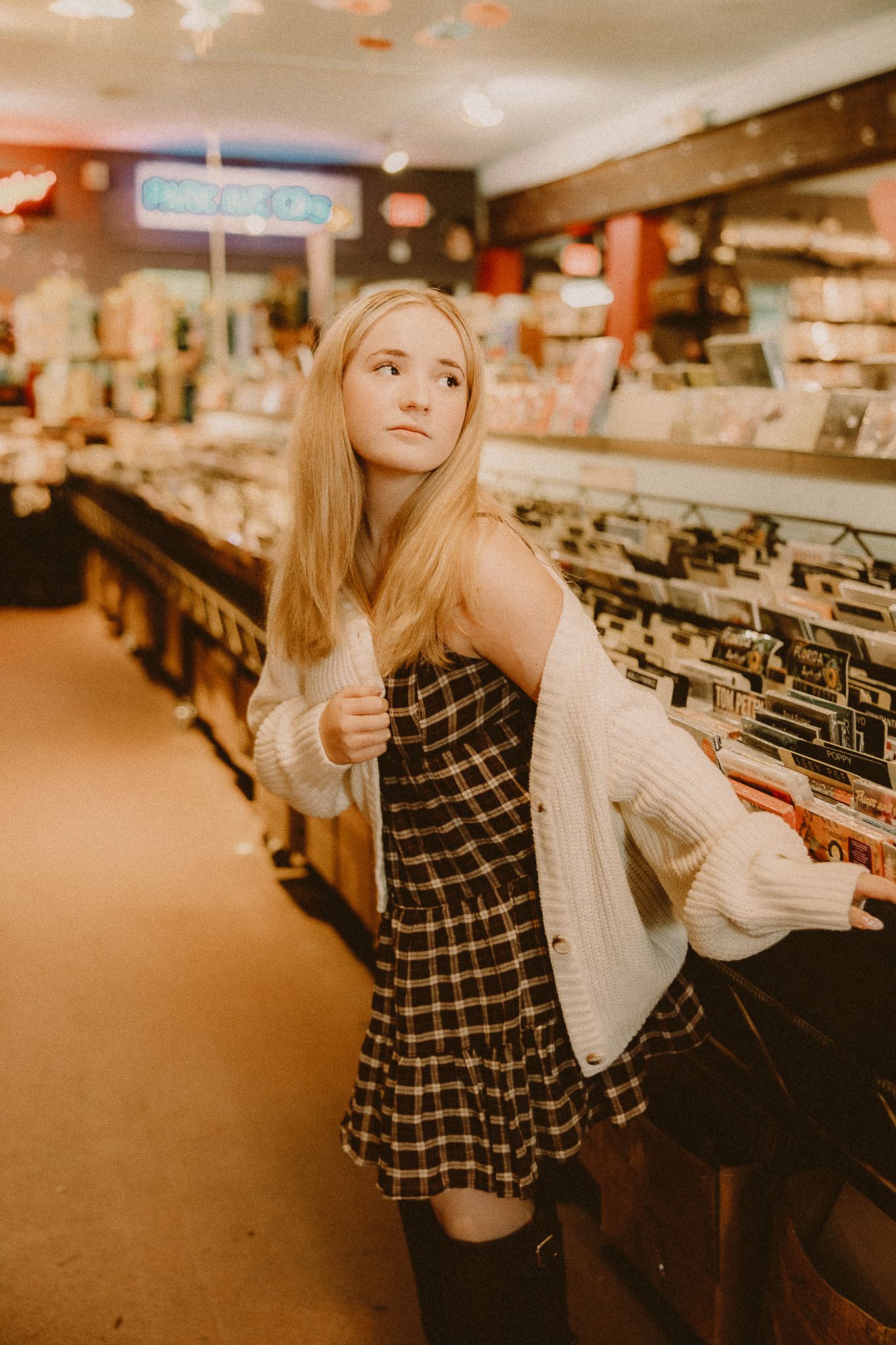Young woman with long blonde hair in a plaid dress and cream cardigan looks over her shoulder in a vintage record store.