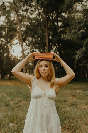 Young woman in a white sundress balances a stack of books on her head while sticking out her tongue in a sunlit forest clearing.