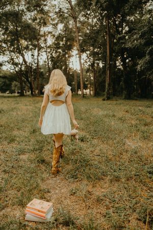 Woman in a white dress and brown cowboy boots walks away through a grassy woodland field, leaving a stack of books behind her.