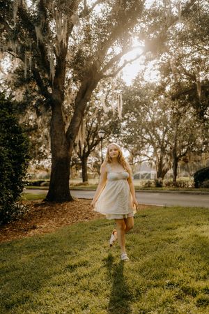 Blonde woman in a white sundress walks across a grassy lawn beneath mossy oak trees as golden sunlight filters through the branches.