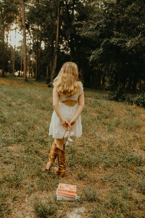 Woman in a white dress and cowboy boots stands with her back to the camera in a grassy forest, holding a stuffed toy near a stack of books.