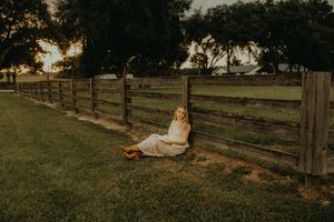 Woman in a white dress sits on the grass leaning against a wooden fence at golden hour with trees in the background.