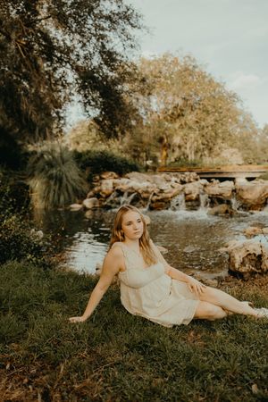 Woman in a cream dress sits on grassy ground beside a small rocky waterfall and creek, surrounded by trees at golden hour.
