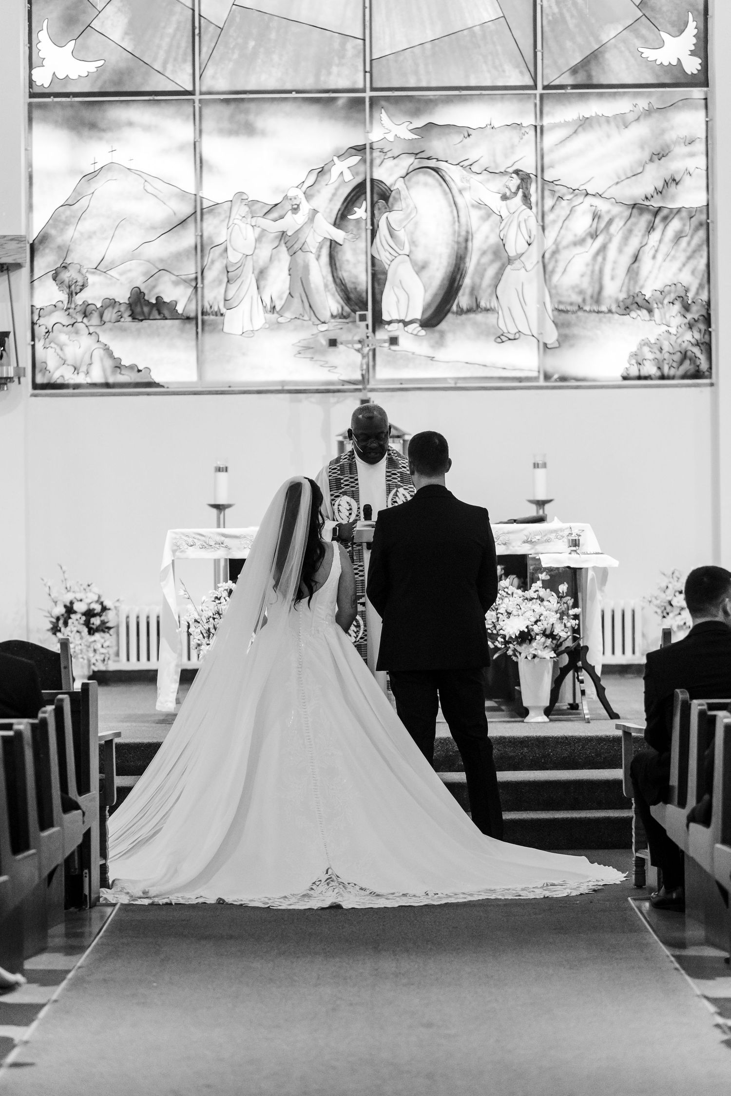 Black and white photo of a bride in a long gown and a groom facing a priest at the church altar during a wedding ceremony.