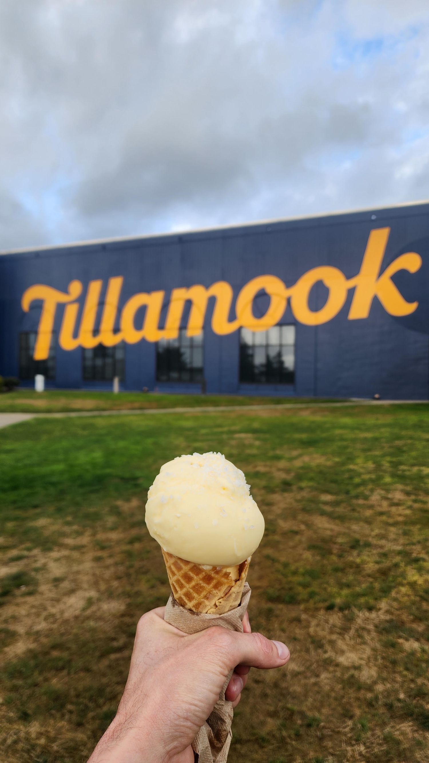 Hand holding a vanilla ice cream waffle cone in front of the Tillamook Creamery building with yellow logo signage. Butter dipped cone