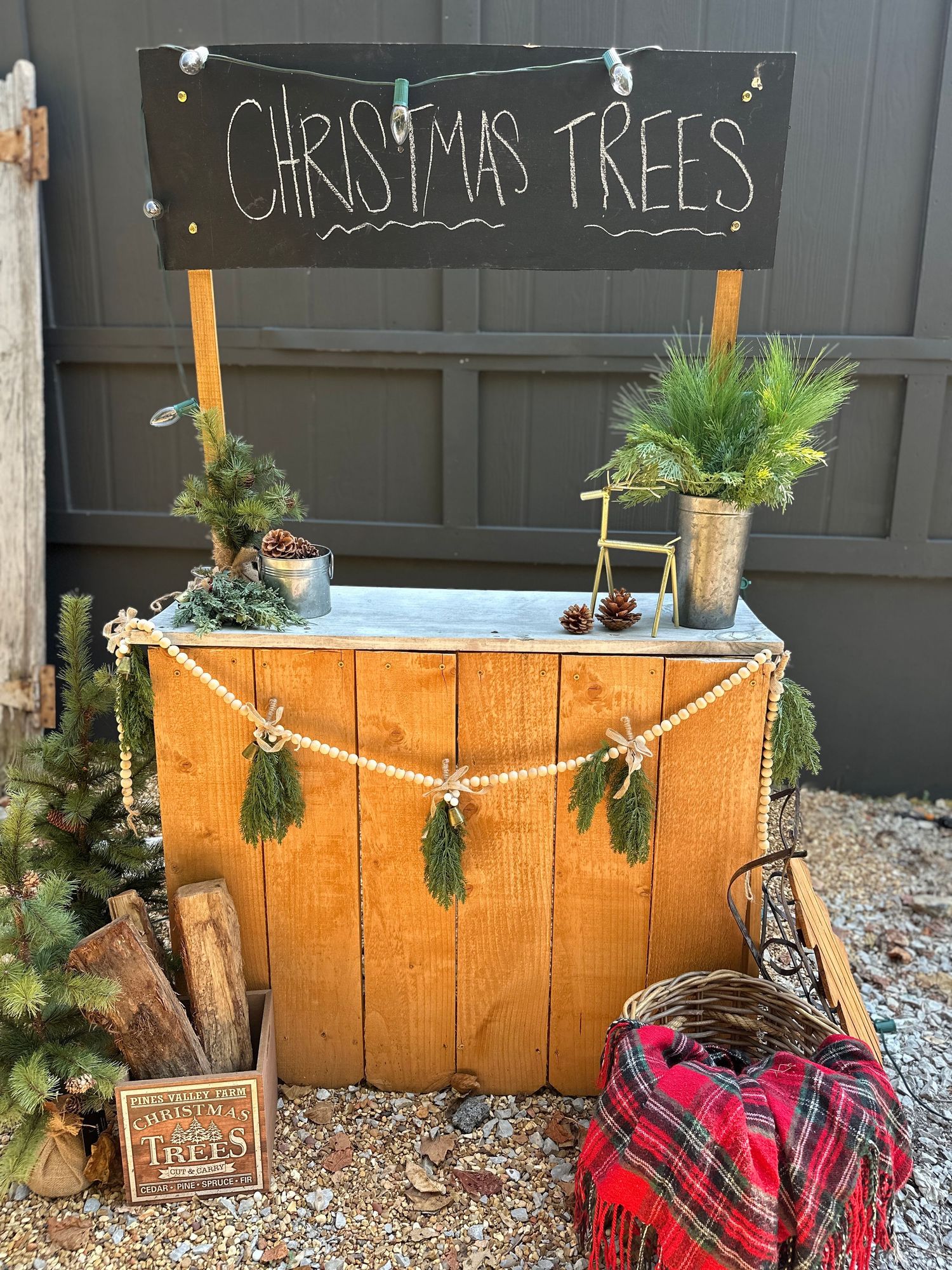 Rustic wooden Christmas tree stand with chalkboard sign, pine garland, and holiday decor against a gray garage door.