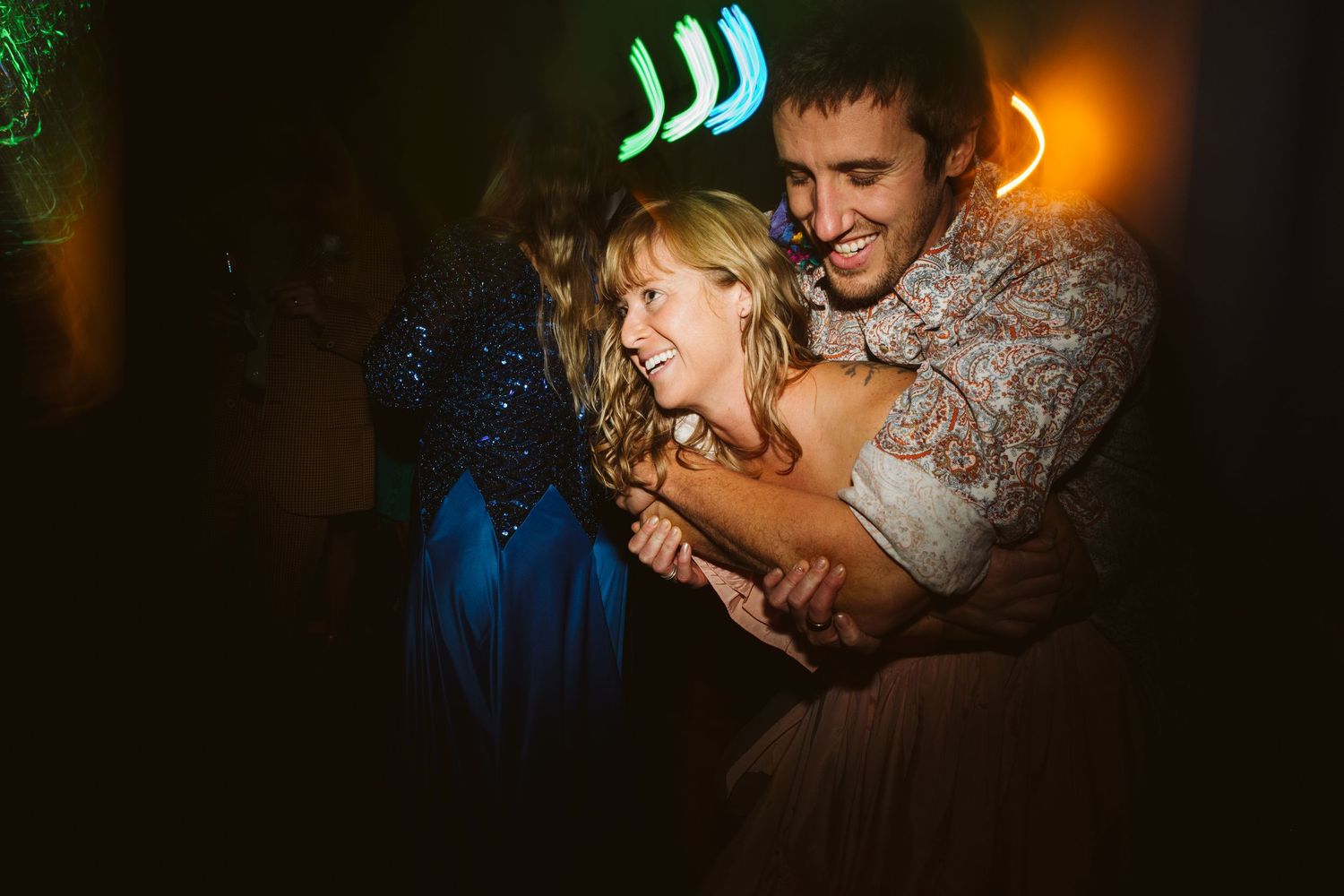 Joyful couple dancing and laughing together at a wedding reception under colorful neon lights.
