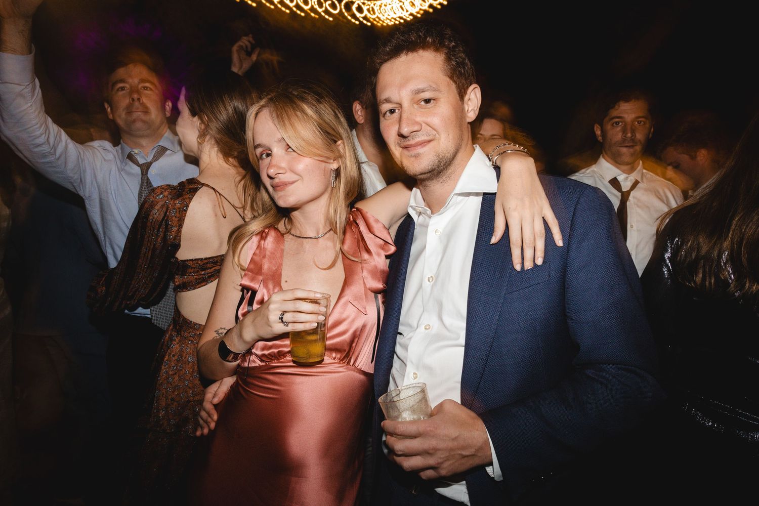 Couple posing together at a lively party, woman in pink dress holding a drink, man in blue suit smiling.