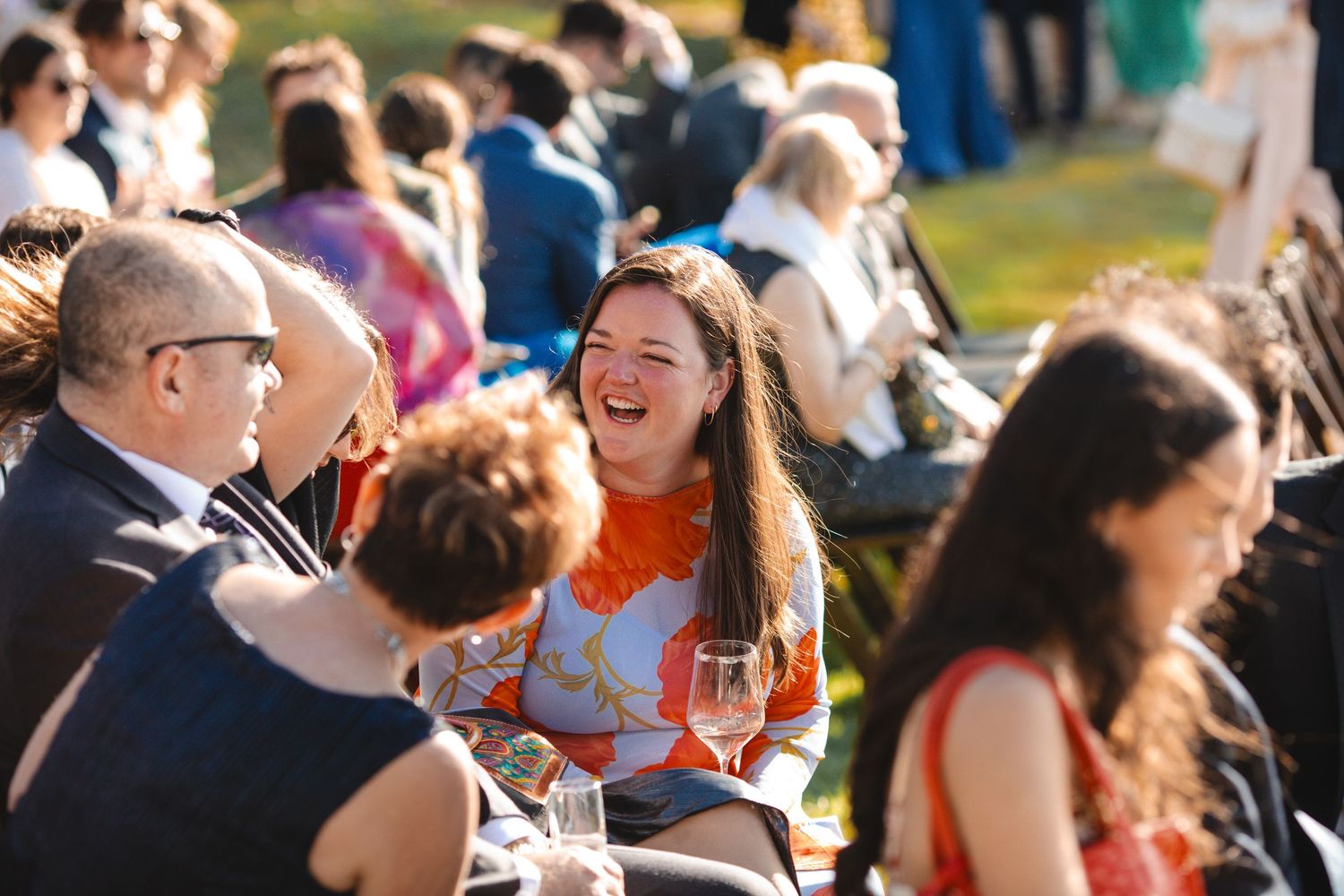 A woman laughs joyfully at an outdoor social gathering, holding a wine glass, surrounded by elegantly dressed guests.