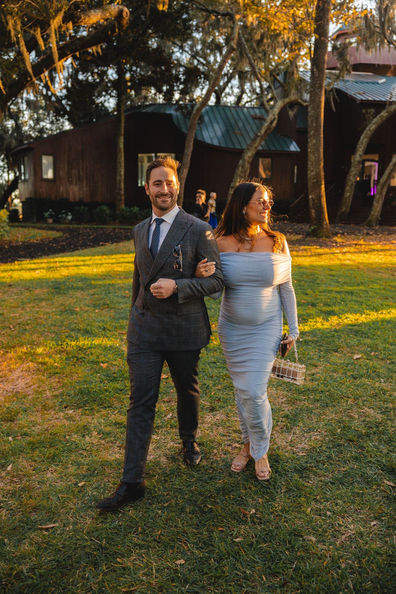 Couple in formal attire walking outdoors at golden hour near rustic cabins, woman in white dress, man in gray suit.