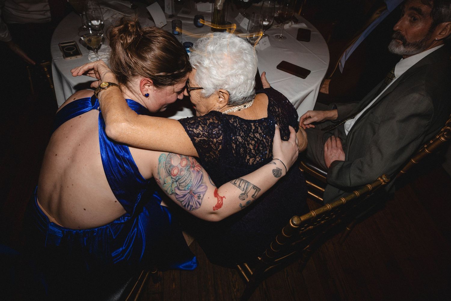 Tattooed woman in blue dress embracing elderly woman in black lace at a formal event.