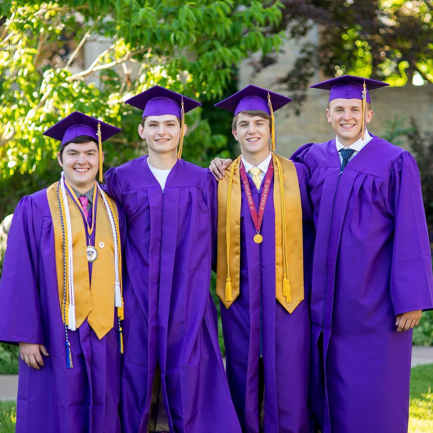 Four male high school graduates in purple cap and gown smiling together outdoors after ceremony.