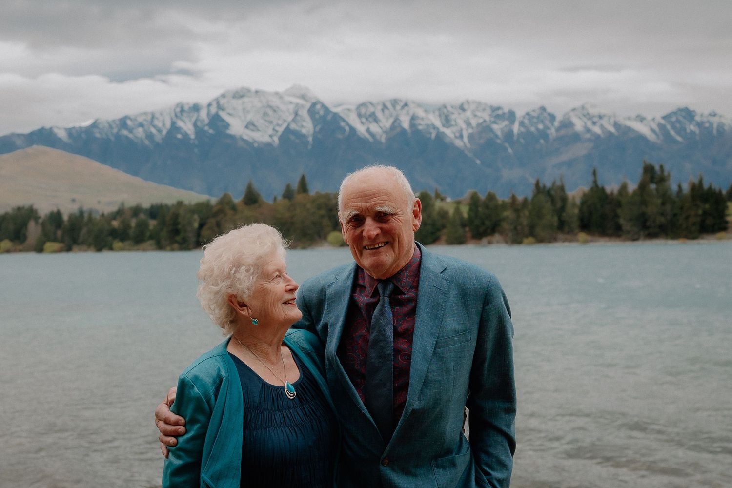 Elderly couple in teal jackets smiling and embracing beside a calm lake with snow-capped mountains and cloudy sky behind them.
