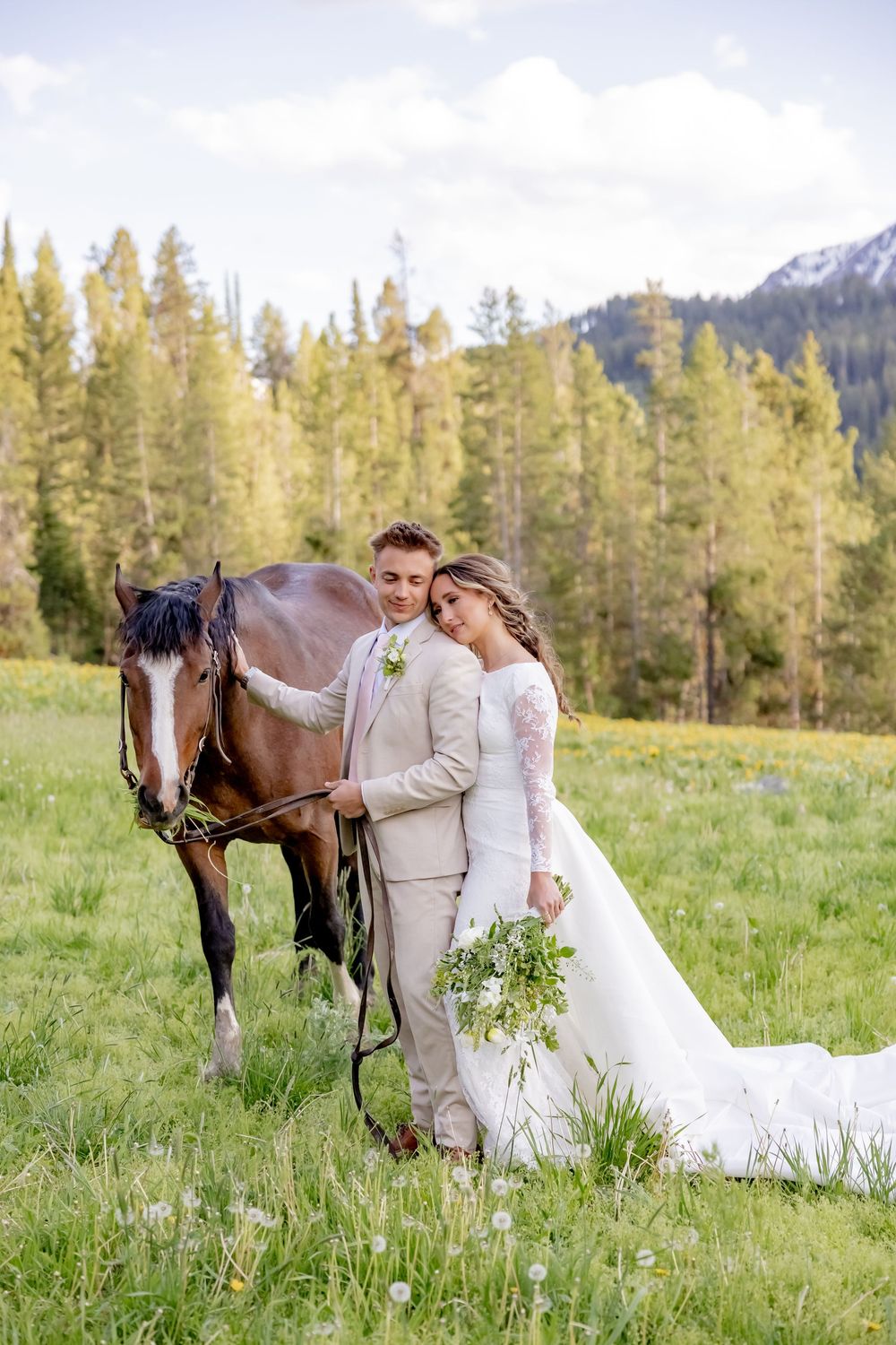Bride in a long-sleeve lace gown and groom in a tan suit stand with a brown horse in a mountain meadow, holding a green bouquet.