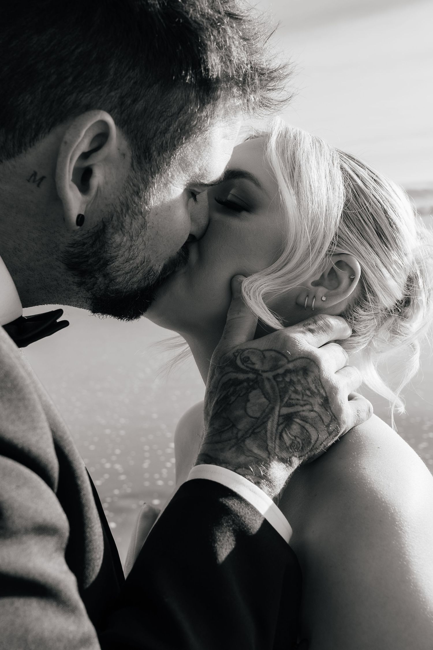 Black and white photo of a bearded groom with an ear stud kissing his bride, his tattooed hand gently cradling her face.