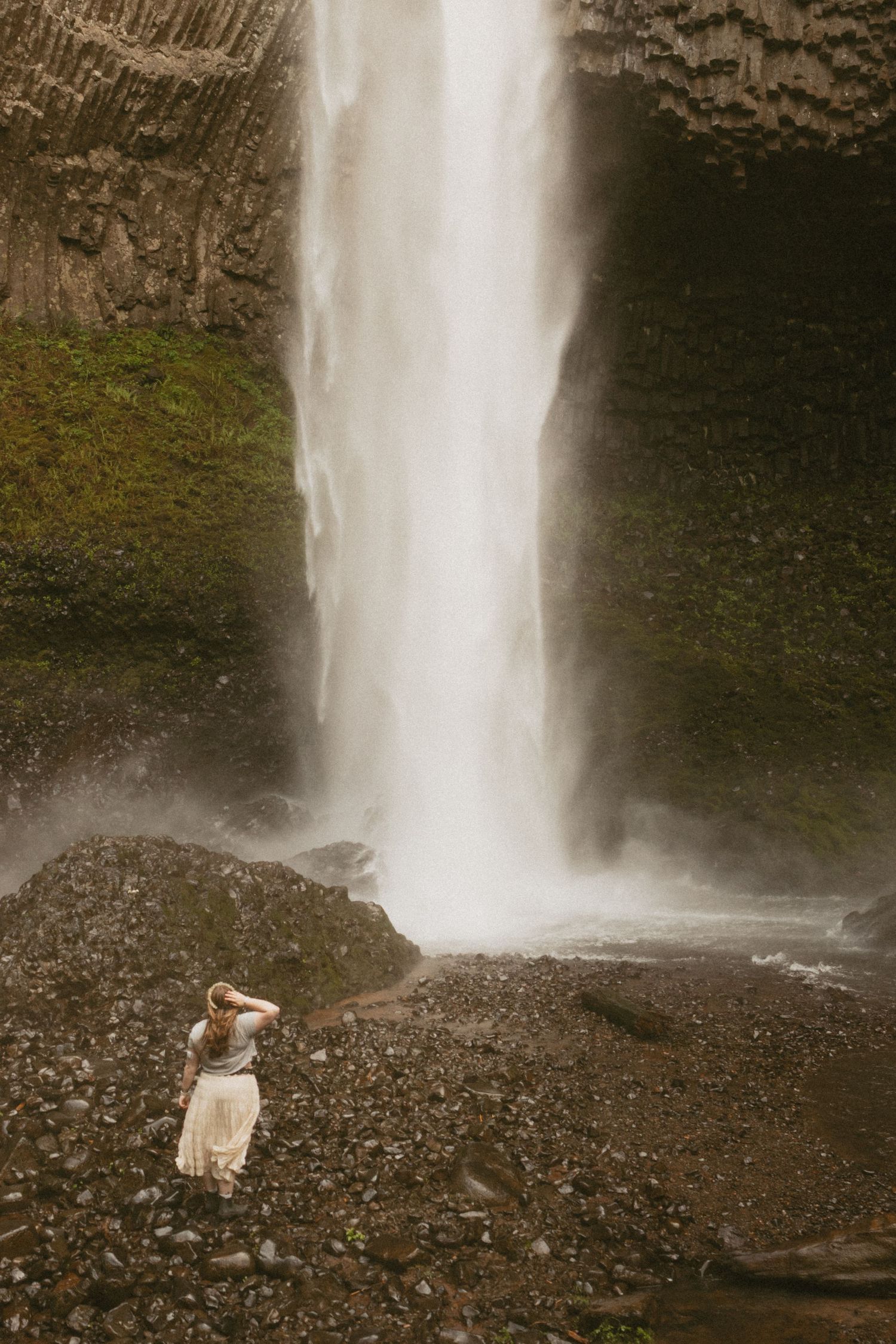 Woman in a white skirt stands on a rocky shore gazing up at a tall waterfall cascading down a mossy basalt cliff.