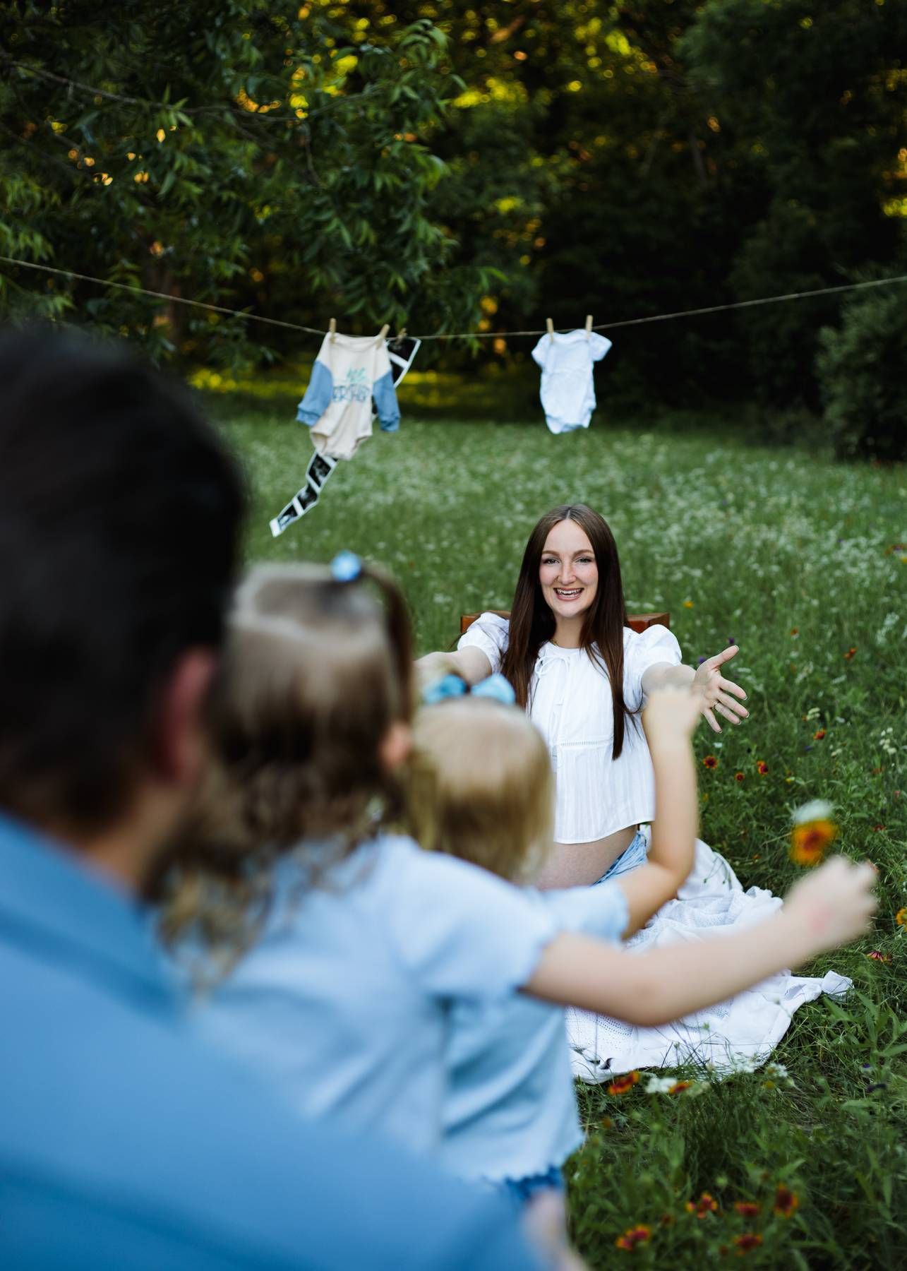 Smiling pregnant mother sits in a meadow with baby clothes hanging on a line as her family reaches toward her.