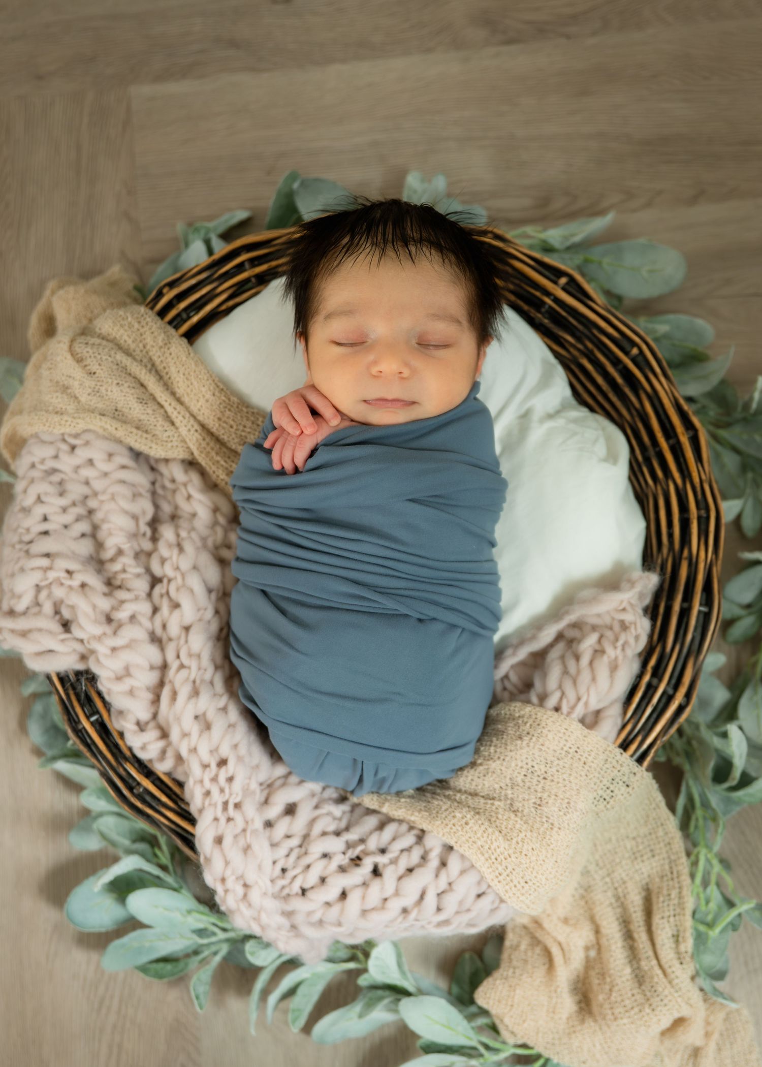 Newborn baby swaddled in blue wrap sleeping peacefully in a wicker basket with knit blankets and greenery.