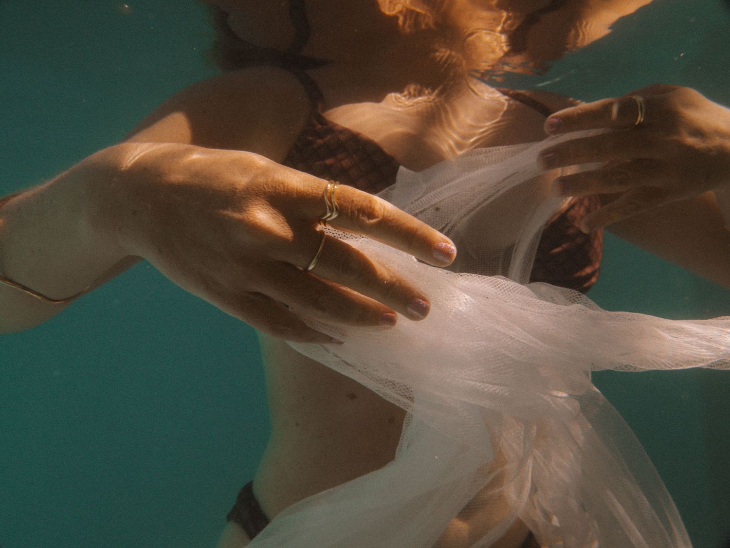 Person wearing gold rings holding white fabric underwater in turquoise pool, underwater fashion photography in Newcastle NSW