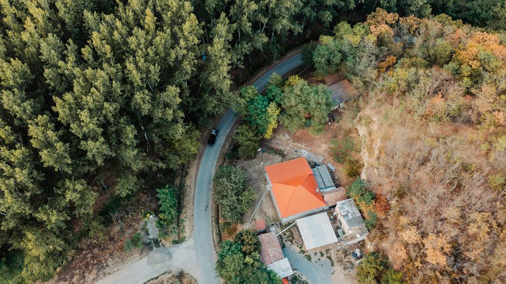 Aerial view of a winding road through dense forest with a small house featuring an orange roof.