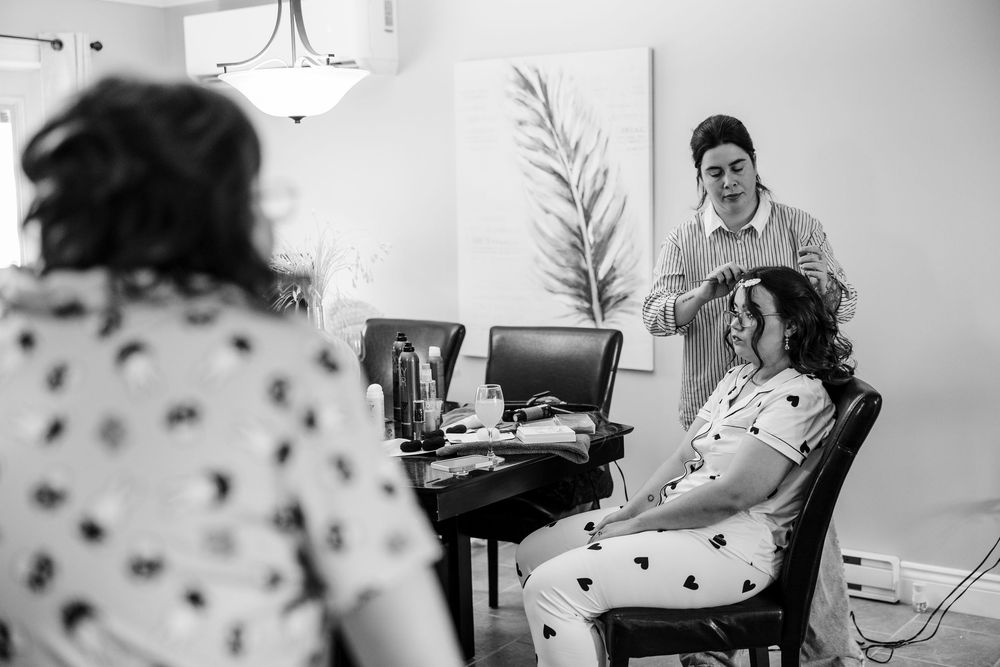 A hairstylist works on a seated woman's hair in a bright room while another woman watches in a black and white photo.