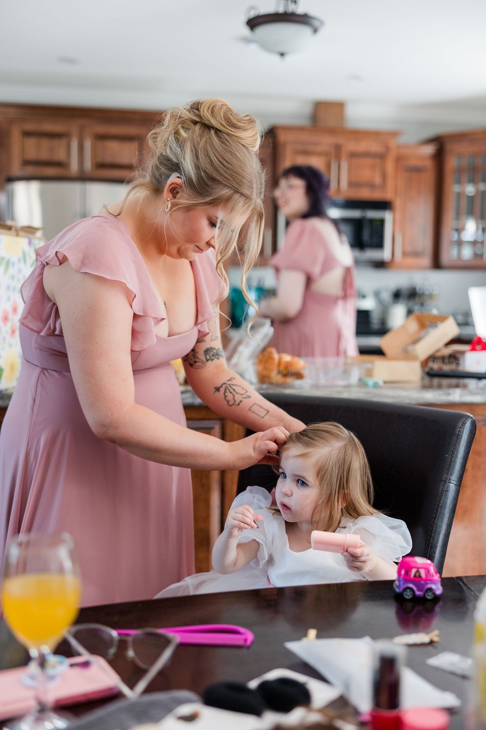 Bridesmaid in pink dress helps young flower girl at kitchen table during wedding preparations.