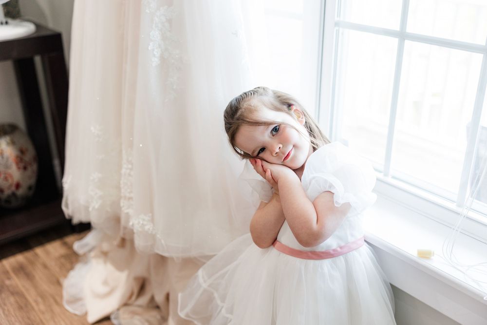 Young flower girl in white dress with pink sash poses thoughtfully by window near bridal gown.