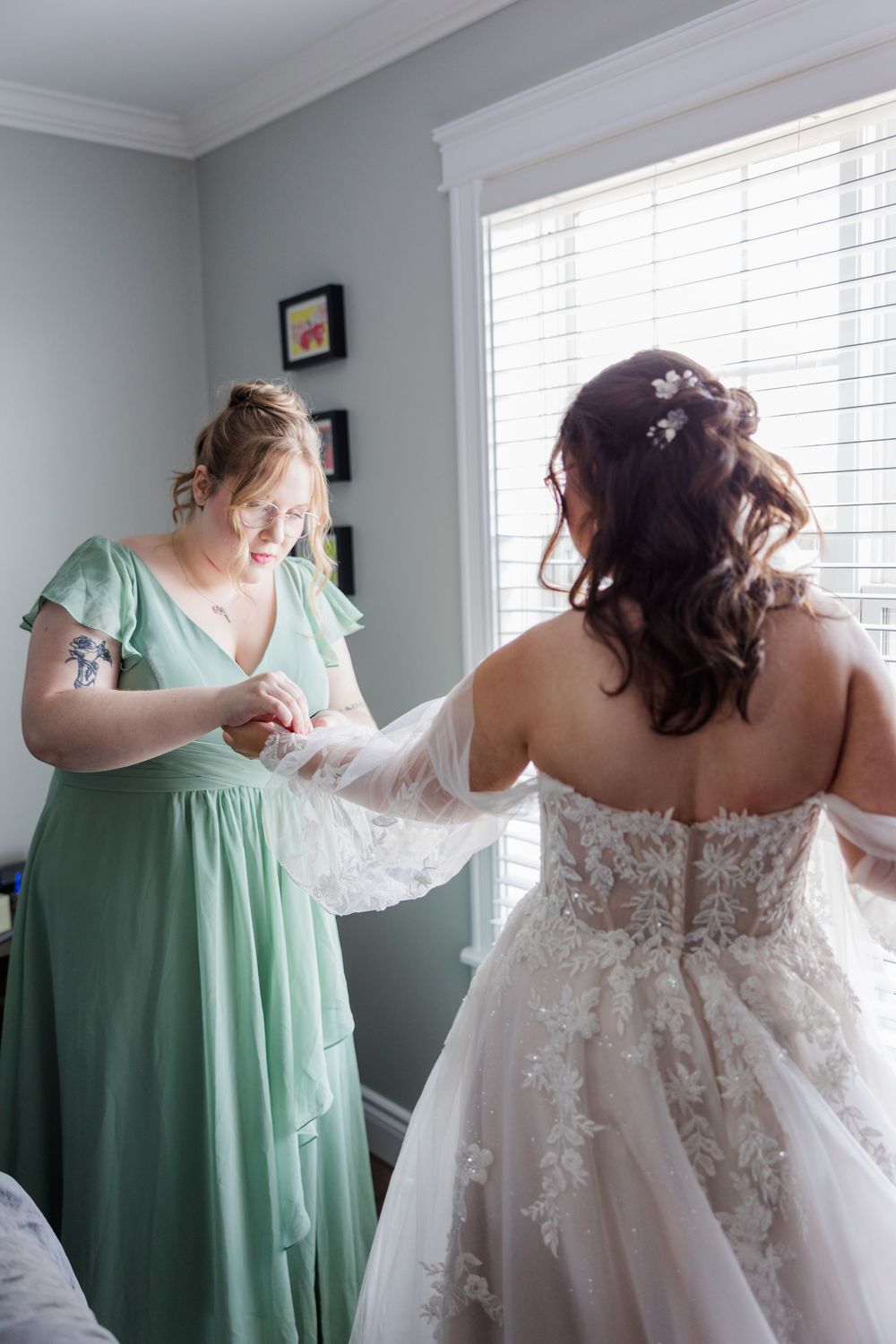 Bridesmaid in sage green dress helps bride button lace wedding gown near bright window.