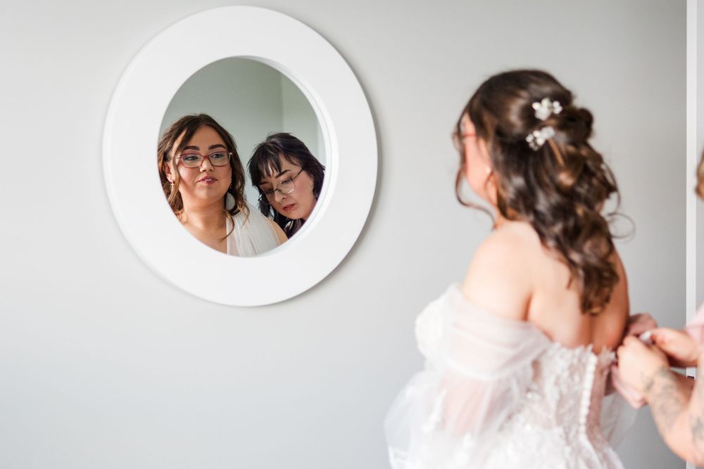 Bride in white gown views reflection with bridesmaid in round mirror during wedding preparations.